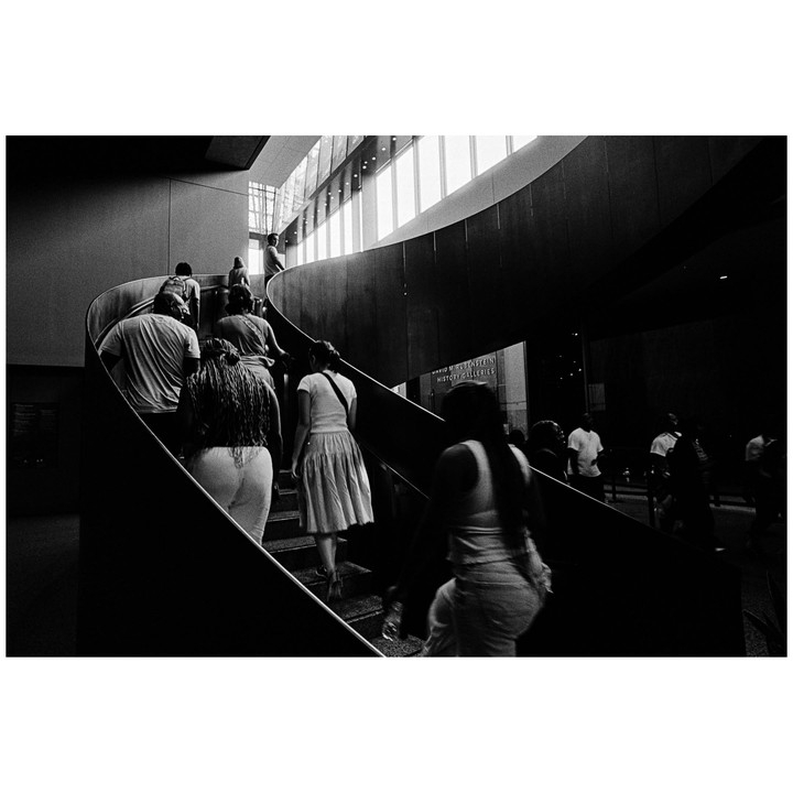 People ascend a spiral staircase in a building with large windows.