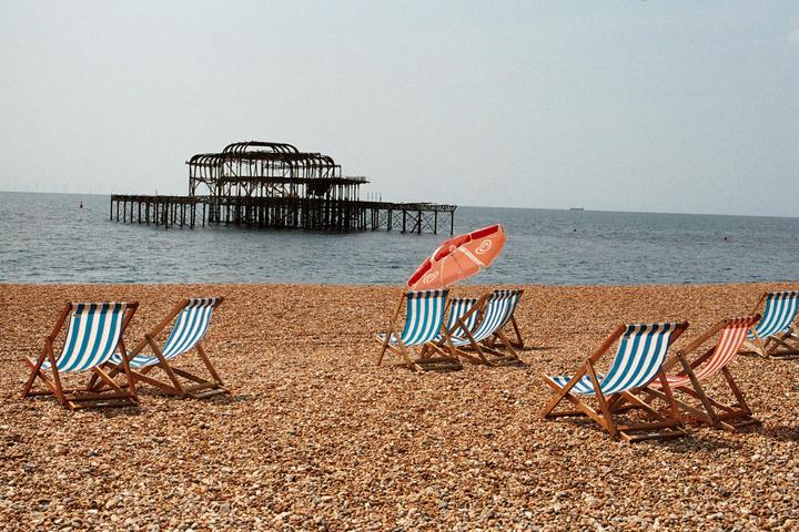 Several striped deck chairs and a red umbrella sit on a pebble beach with a ruined pier in the background.
