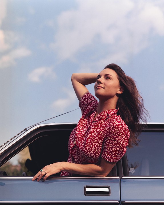 A woman in a red floral shirt leans out of a car window with her arm behind her head, looking up at the sky.