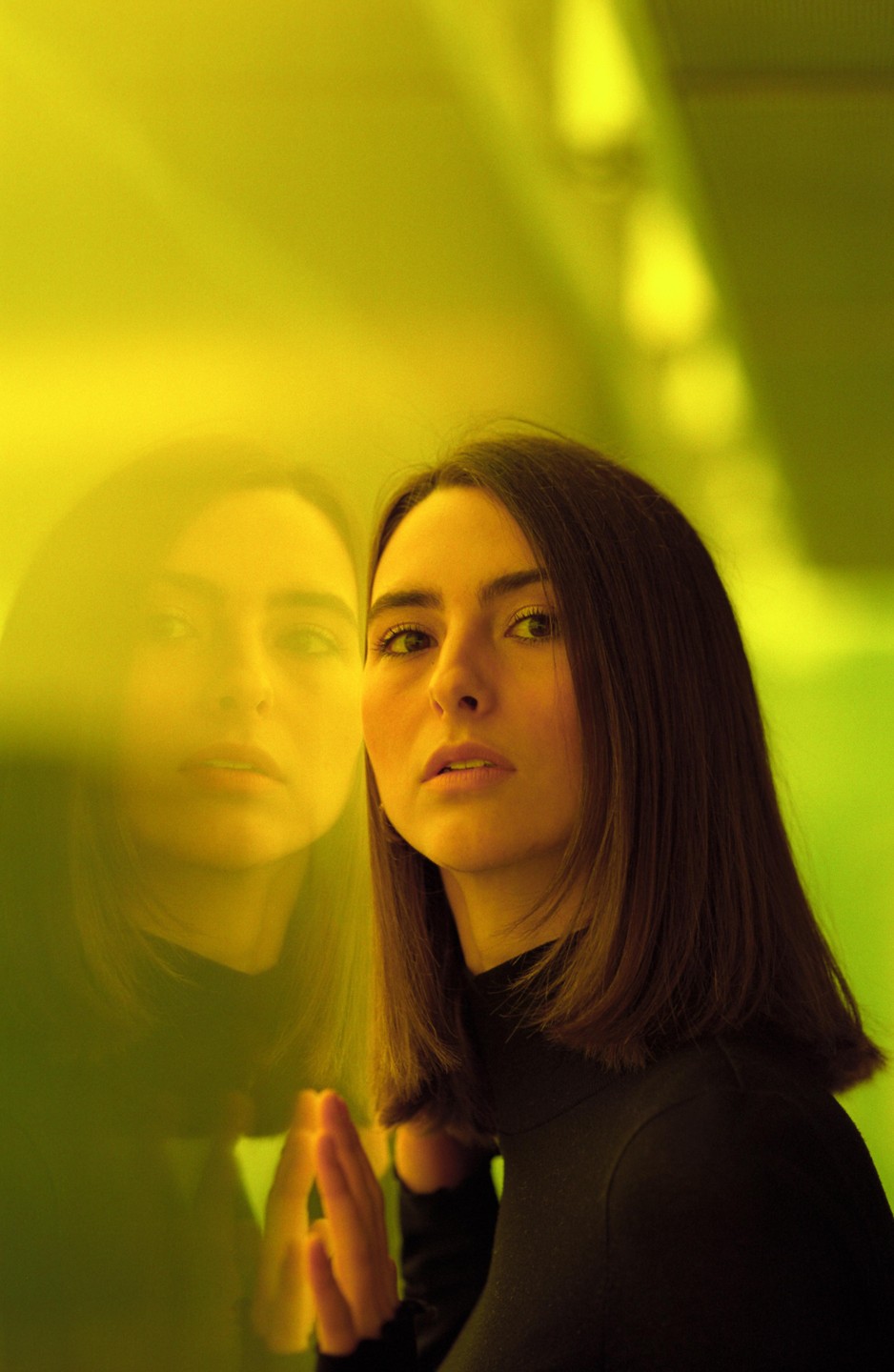 A woman with dark hair looks at the viewer, her reflection visible on a yellow surface.