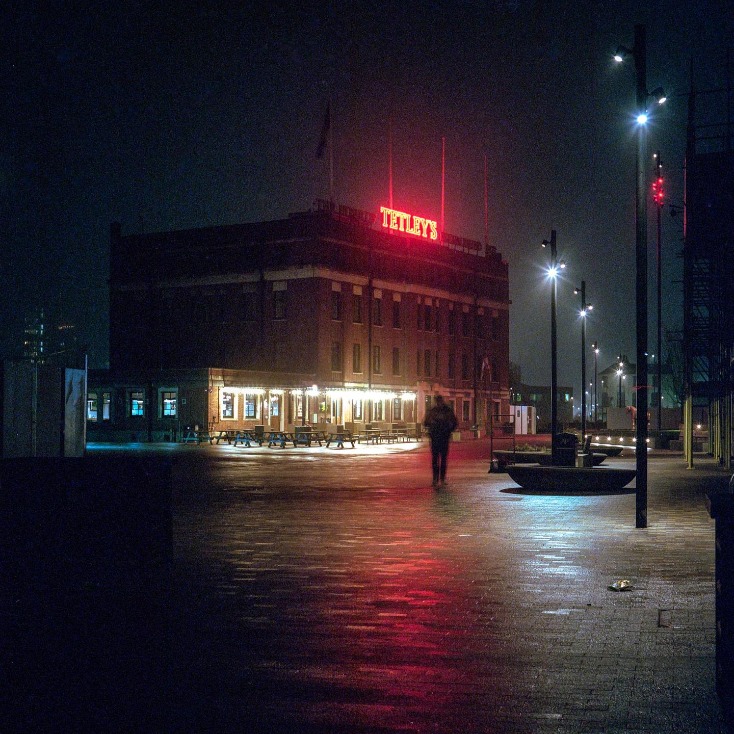 A person walks on a wet street at night with a building and neon sign in the background.