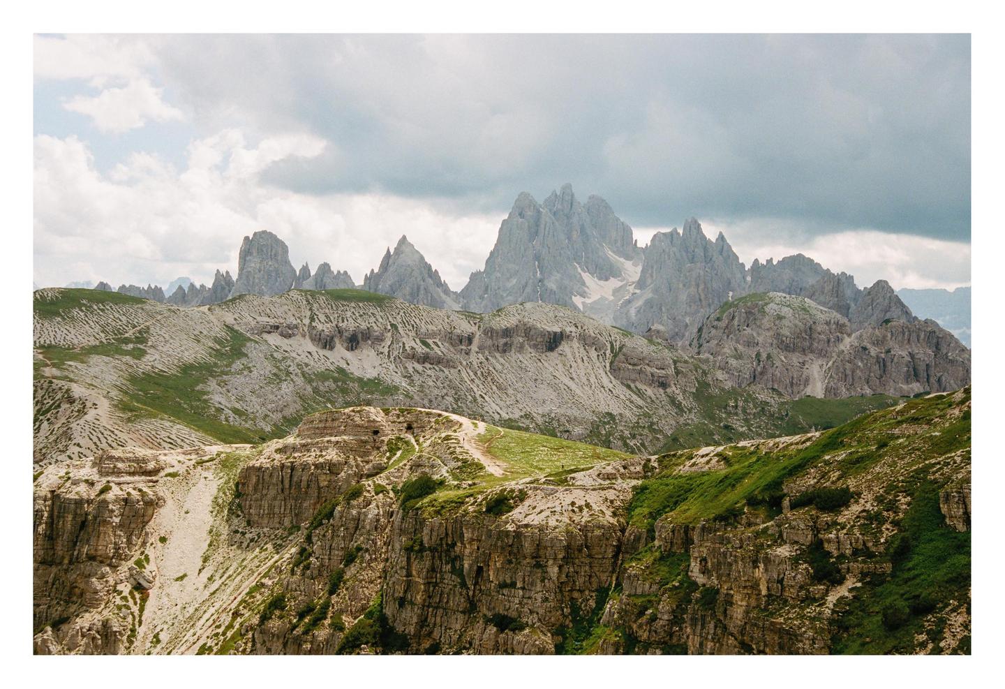 A hiking trail winds through a mountainous landscape with jagged peaks under a cloudy sky.
