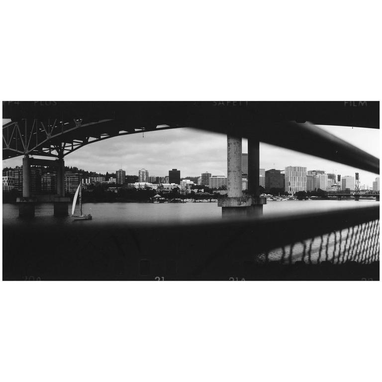 A panoramic black and white image shows a cityscape across a body of water, framed by the underside of a bridge and a sailboat in the foreground.