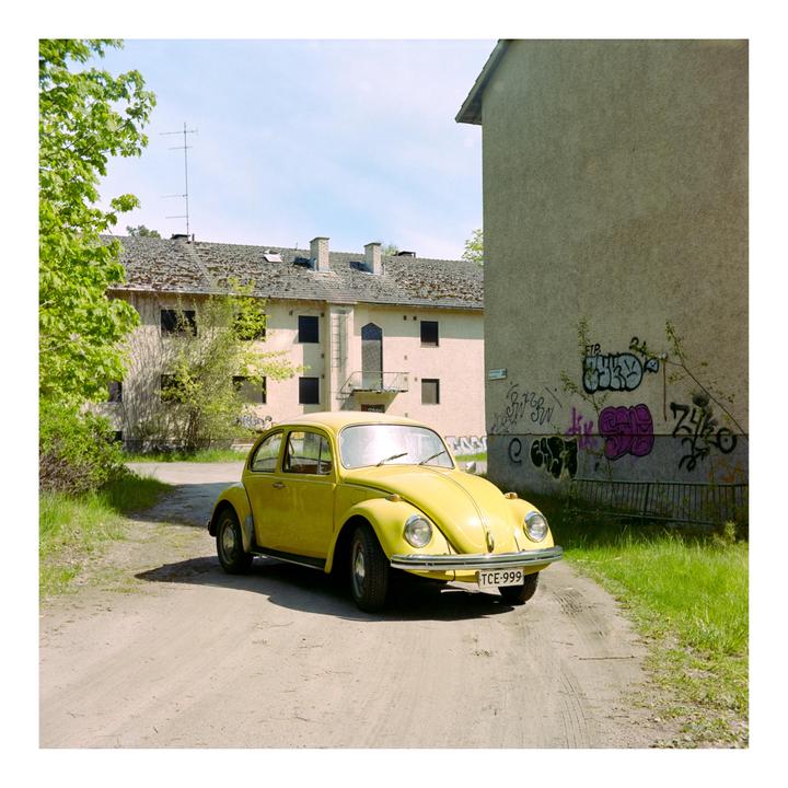 A yellow volkswagen beetle is parked on a dirt road next to buildings with graffiti.