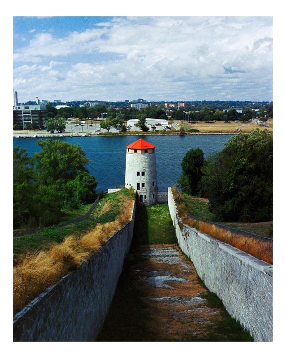 A stone tower with a red roof stands by a body of water, with a city visible in the distance under a cloudy sky.