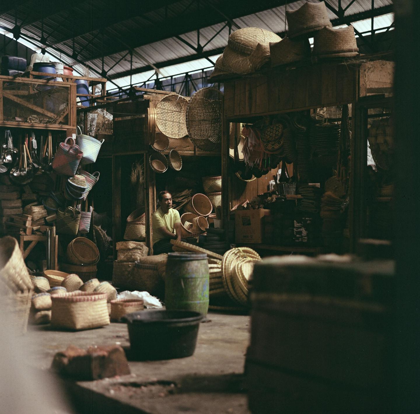 A man sits among many woven baskets and other goods in a market stall.