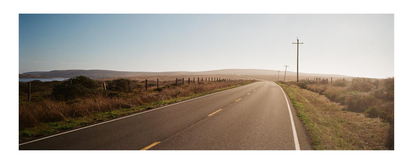 A long, empty road stretches into the distance with hills and a body of water visible under a clear sky.