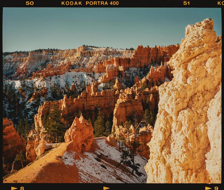 A vast canyon landscape features orange rock formations and scattered snow.