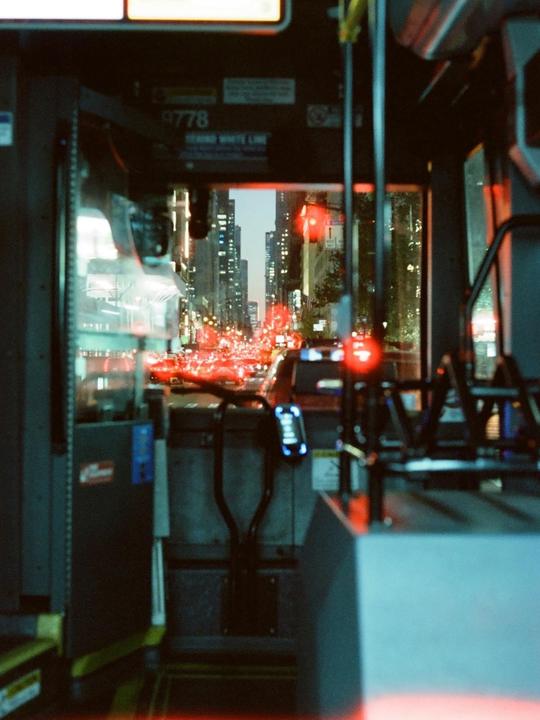 A view from inside a bus shows a city street at night with red traffic lights and tall buildings.