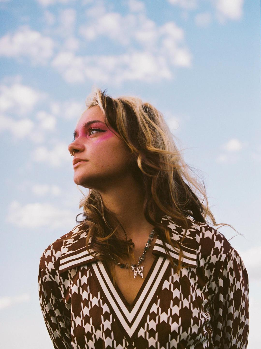 A woman with pink eye makeup and a patterned shirt looks up at a cloudy sky.