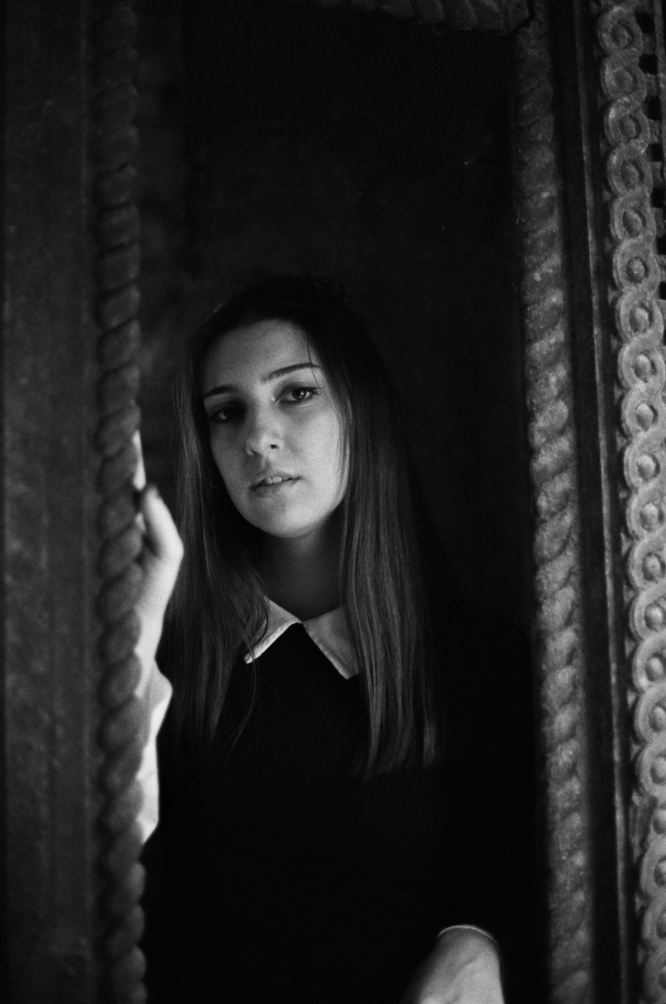 A woman with long hair and a white collar is framed by an ornate doorway.