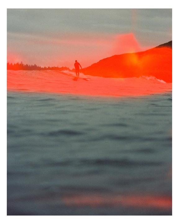 A surfer rides a wave with a bright orange light leak across the water and distant shore.