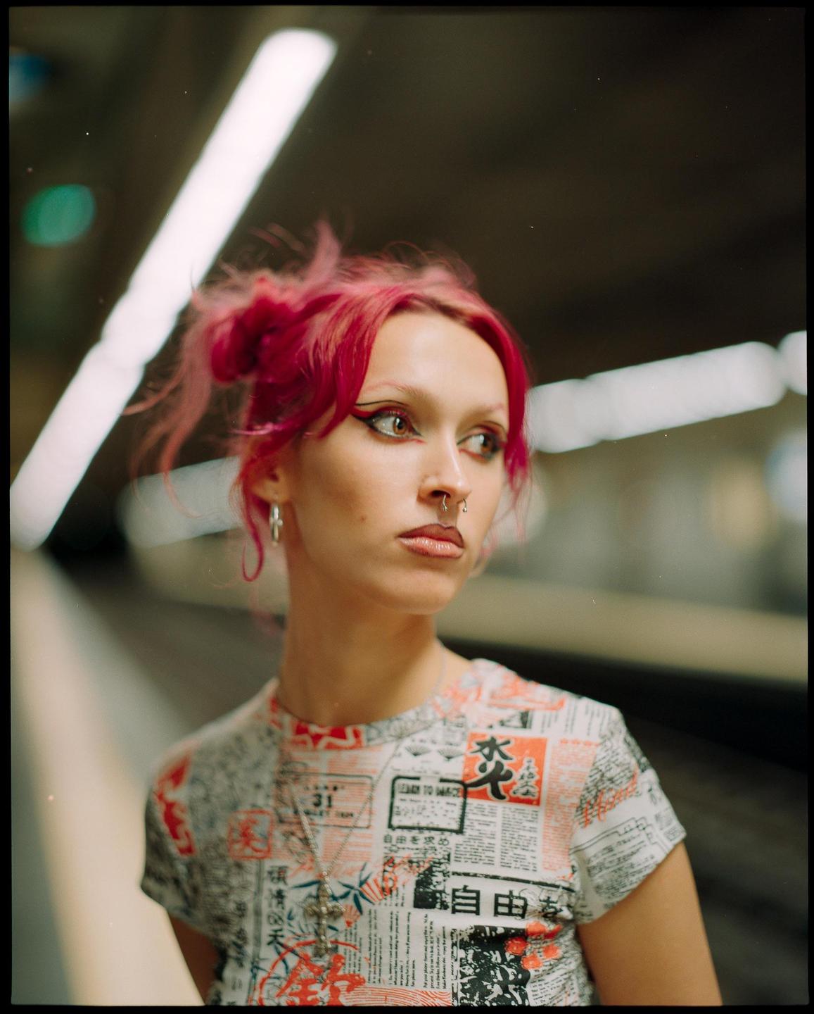 A woman with pink hair and a nose ring looks to the right while standing in a subway station.