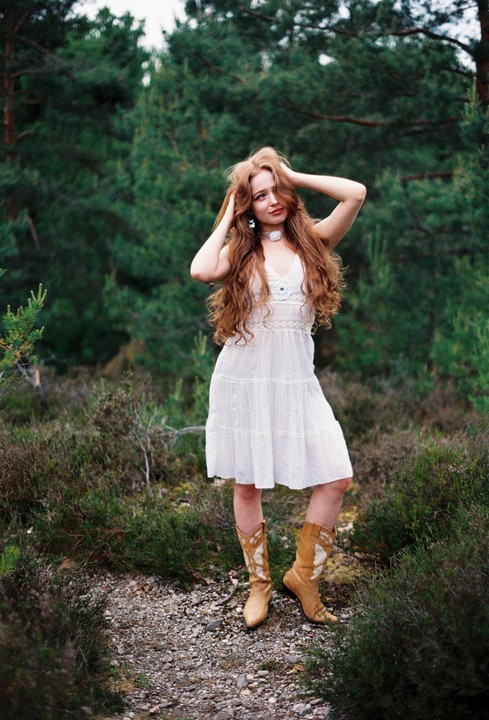 A woman with long, wavy red hair and a white dress stands in a forest.