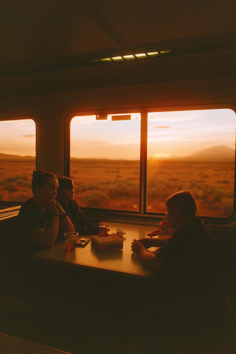 Three people sit at a table inside a train car as the sun sets over a vast landscape outside the window.