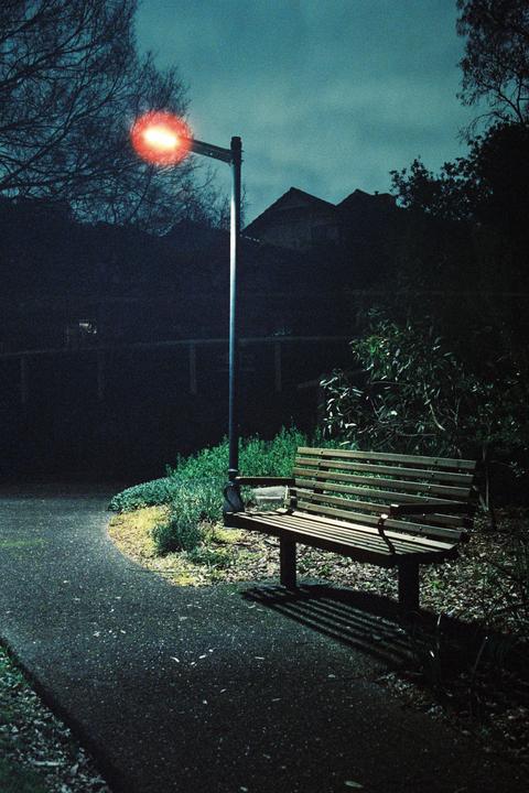 A park bench sits next to a path under a glowing red street light at night.