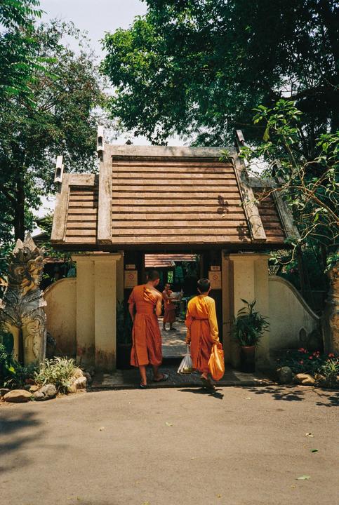 Two monks in orange robes walk towards a temple entrance with a tiled roof, while another monk is visible inside the entrance.