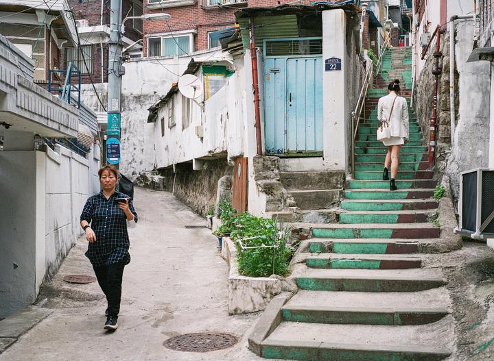A woman walks up a set of stairs with green and brown painted steps while another woman walks on a street below.