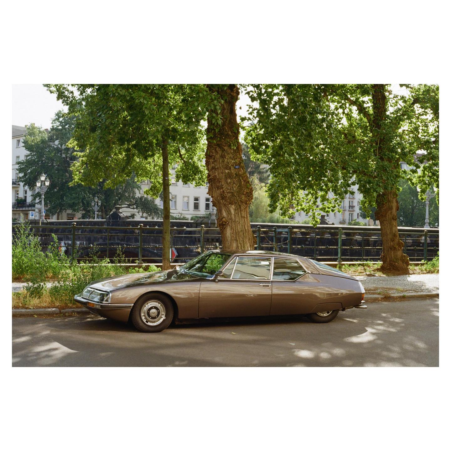 A brown vintage car is parked on a street next to a canal with trees and buildings in the background.