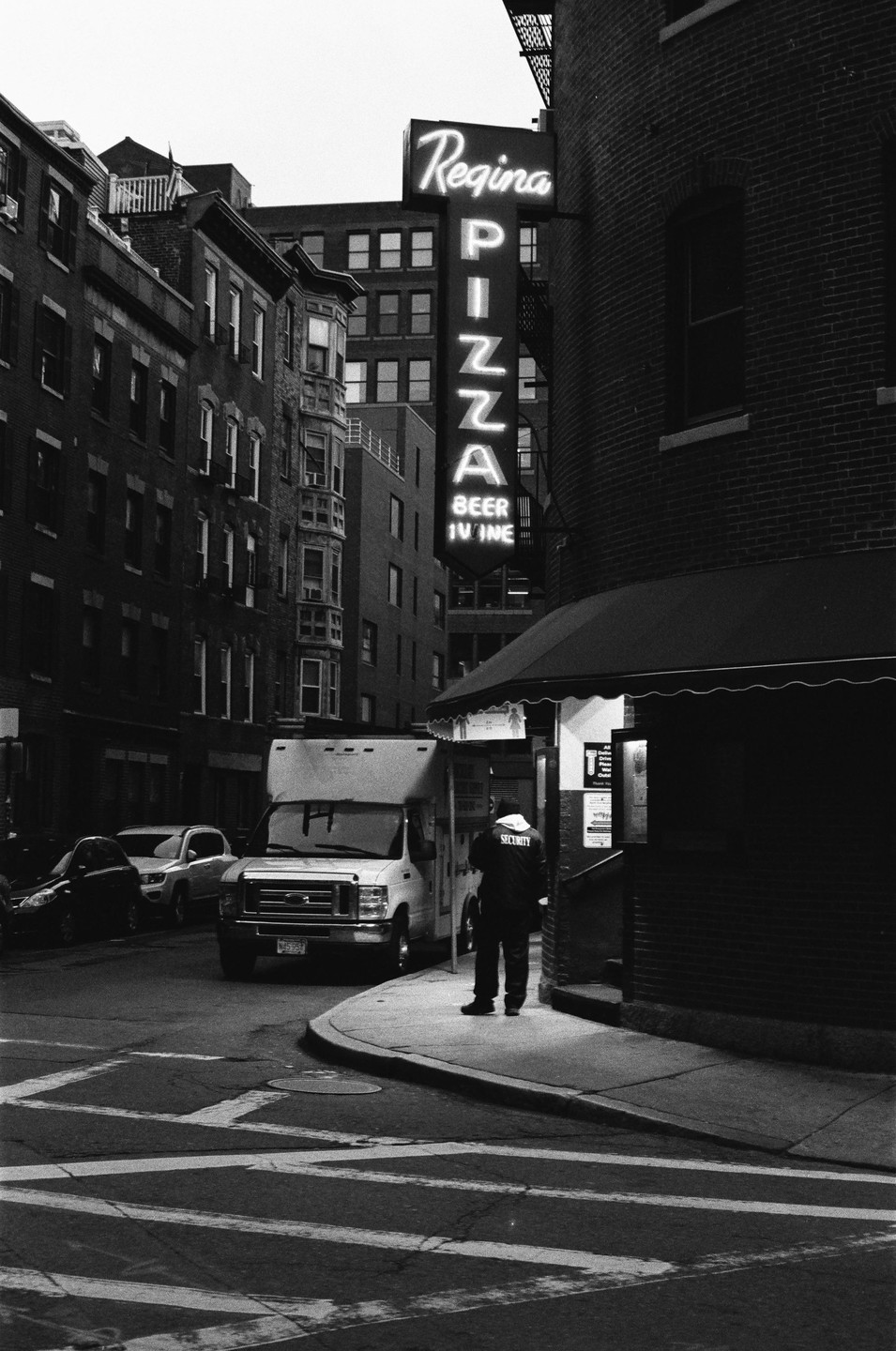 A man stands on a sidewalk next to a white van and a building with a neon "Regina Pizza" sign.