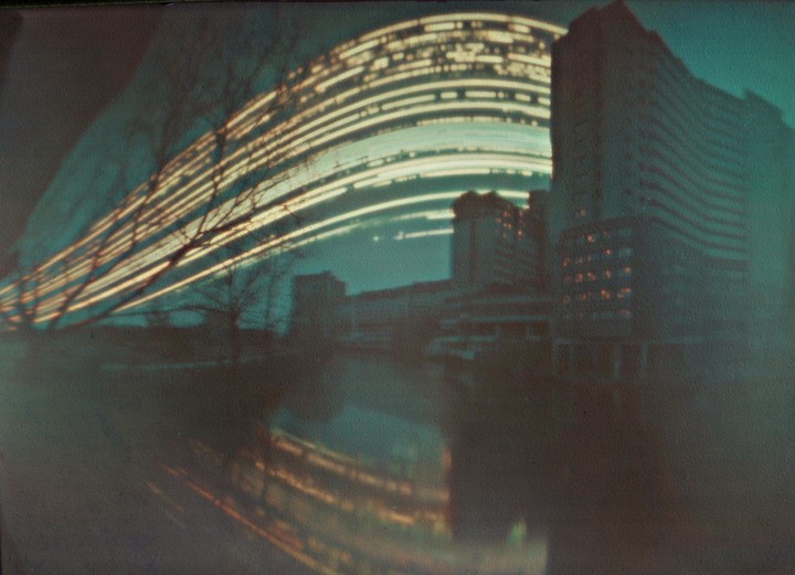 Light trails arc over a brutalist building and its reflection in water.