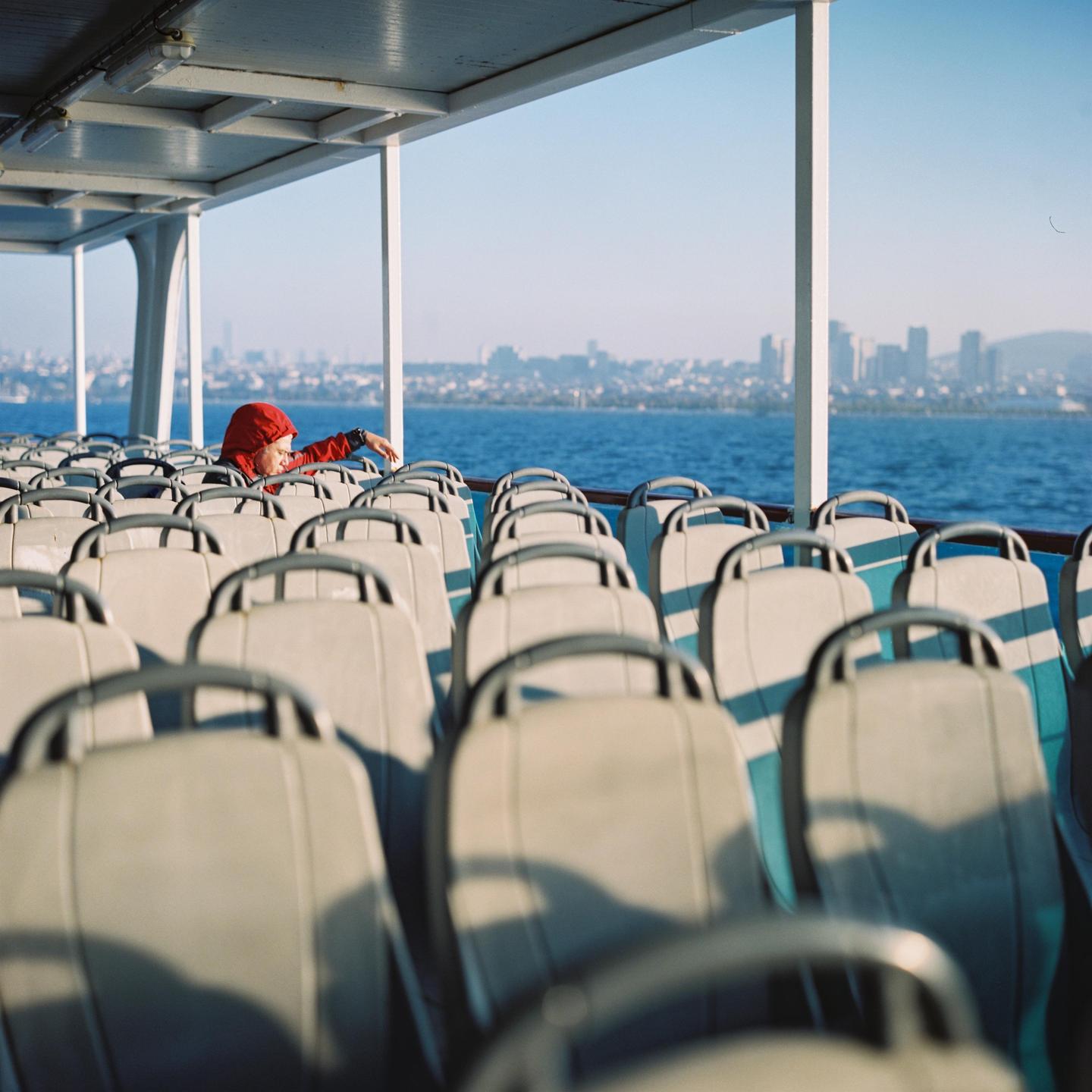 A person in a red jacket sits on a ferry looking out at a city skyline across the water.