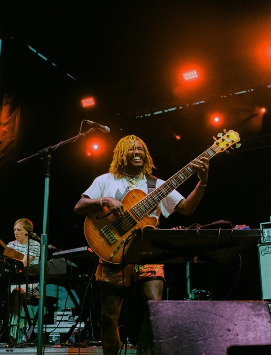 A musician with yellow dreadlocks plays a bass guitar on stage under red lights, with another musician playing a keyboard in the background.