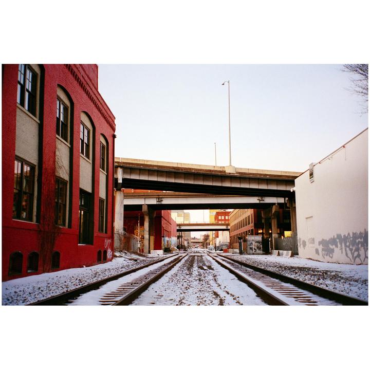 Snow-covered train tracks lead toward a city under an overpass, with a red brick building on the left and a white building on the right.