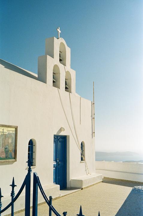 A white church with a blue door and bell tower stands under a clear sky, overlooking a body of water.
