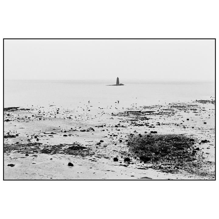 A black and white landscape shows a rocky beach with a lighthouse in the distance and two people walking on the shore.