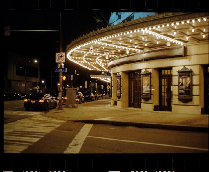 A street corner at night features a brightly lit building with an ornate marquee and cars parked along the curb.