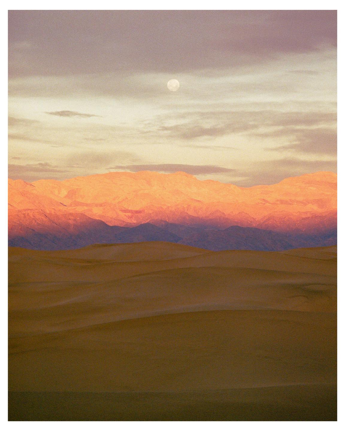 A full moon is visible above mountains and sand dunes at dawn.