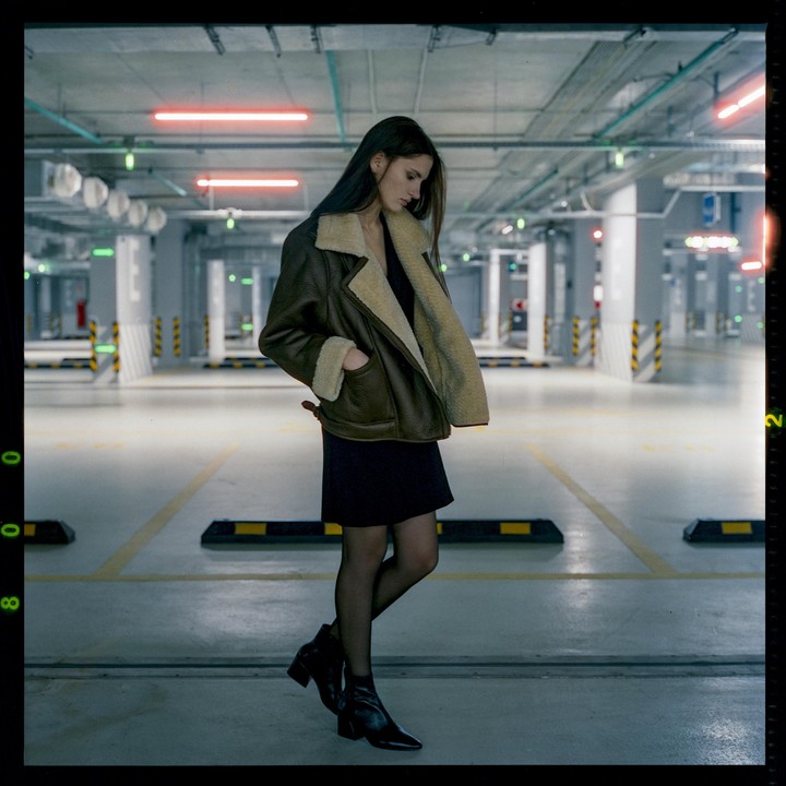 A woman wearing a shearling jacket and a black dress stands in an empty parking garage.