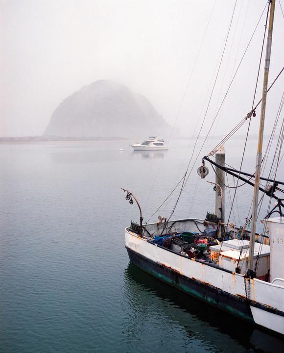 A fishing boat is docked in the foreground with a yacht and a large rock formation in the foggy background.
