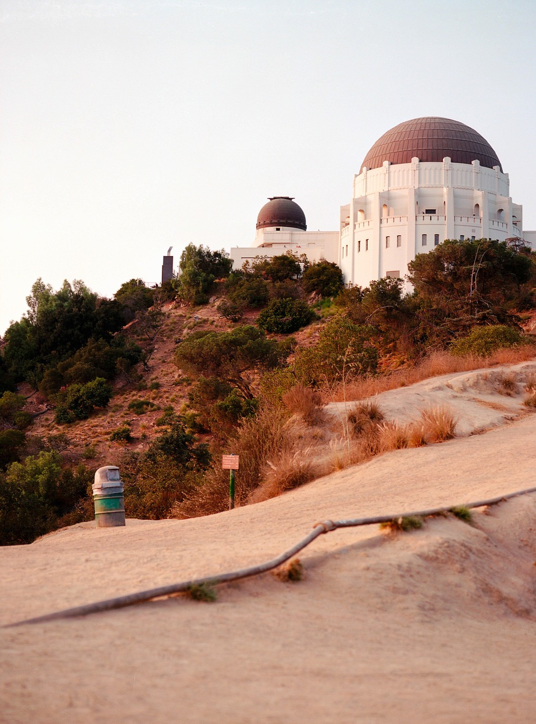 A white observatory building with a large dome sits atop a hill with dry brush and green trees, with a dirt path in the foreground.