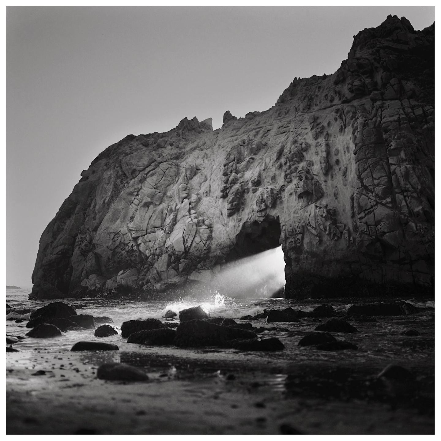 A beam of light shines through a keyhole arch in a large rock formation on a beach with waves crashing on the shore.