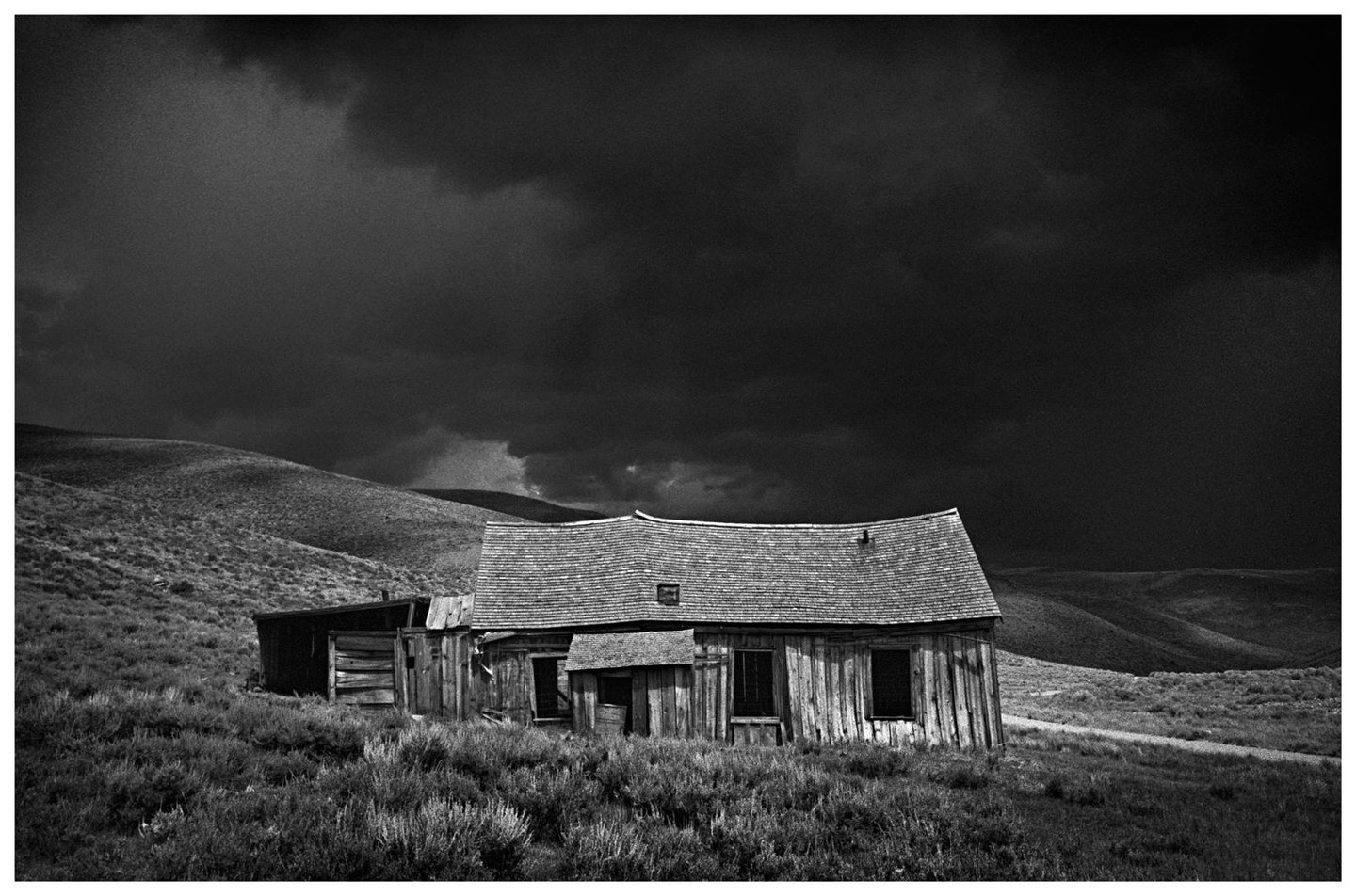 An abandoned wooden house with a curved roof stands in a grassy, hilly landscape under a dark, cloudy sky.