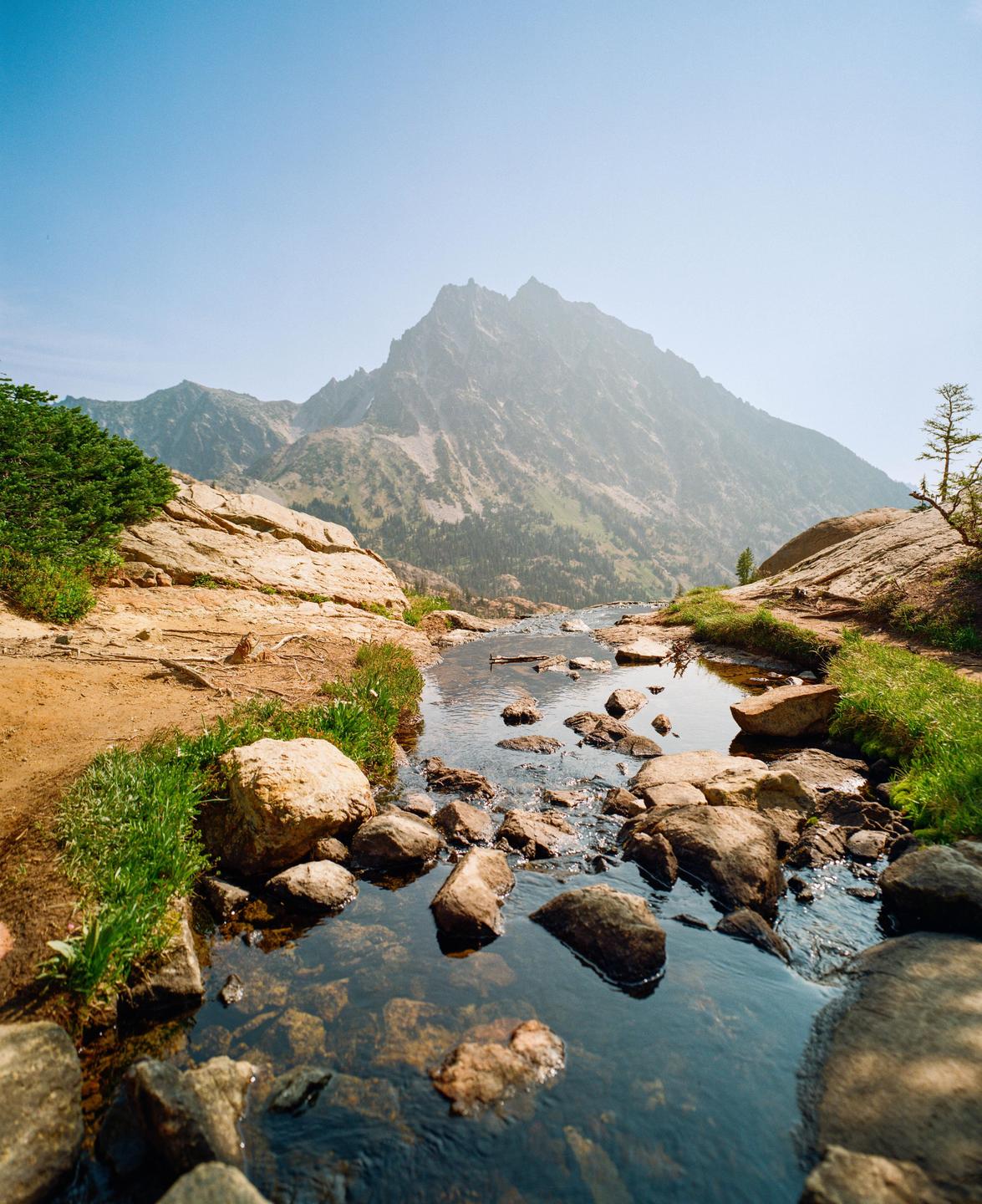 A stream flows over rocks in a mountainous landscape under a clear sky.