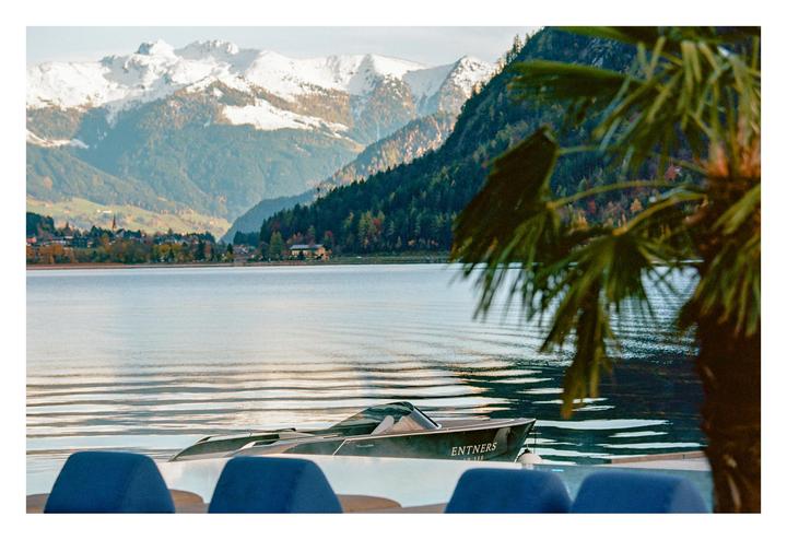 A boat is docked on a lake with mountains and a town in the background, framed by a palm tree.
