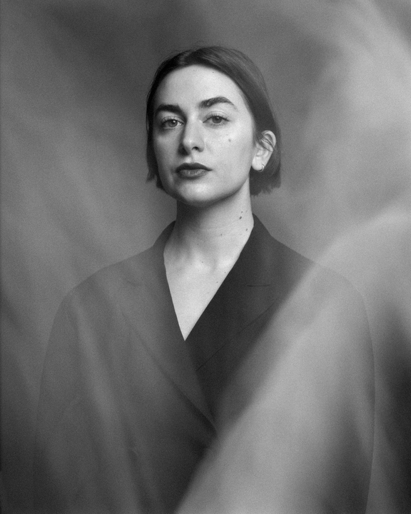 A woman with short hair and a dark jacket looks directly at the viewer.