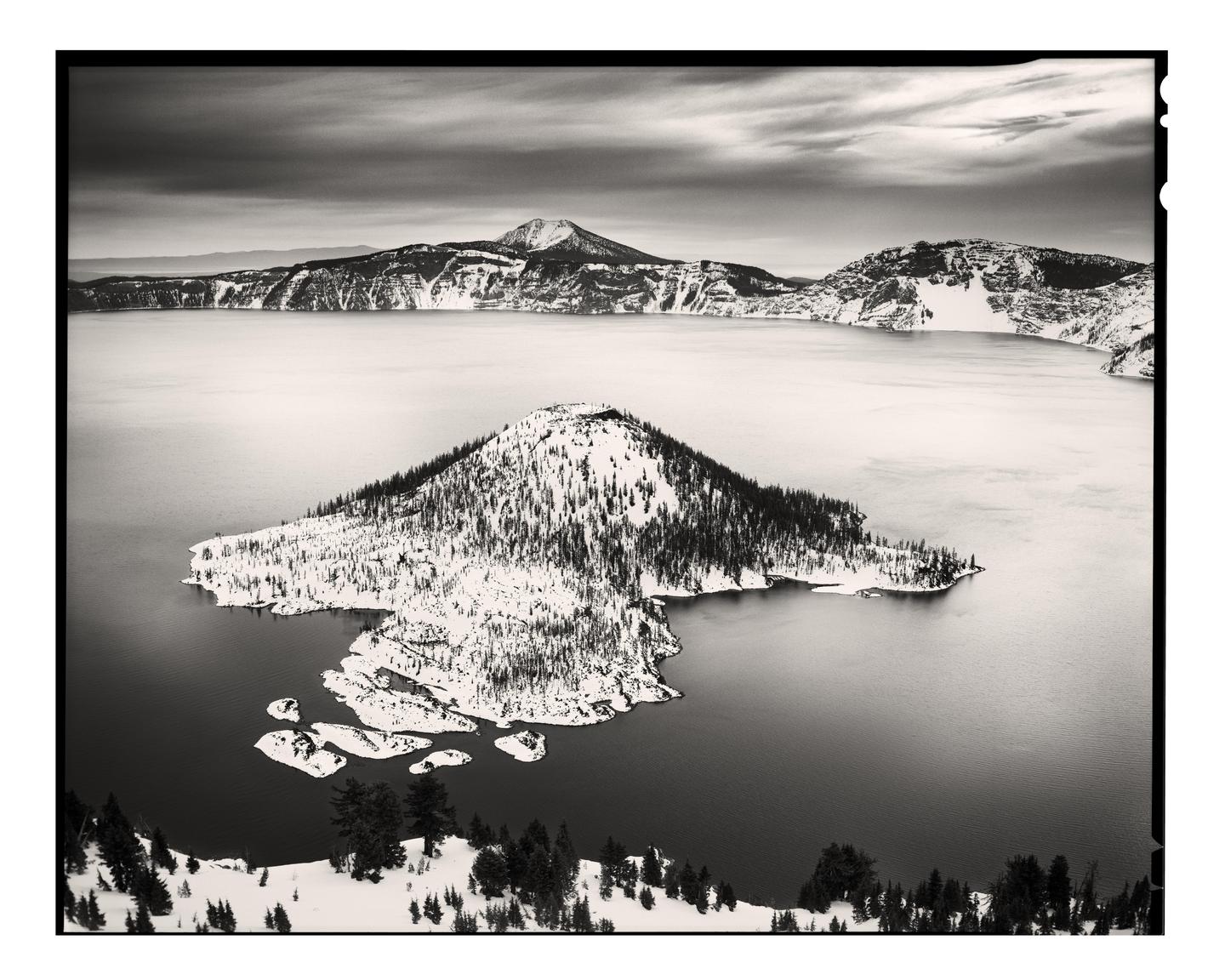 A snow-covered island with trees sits in the middle of a large lake surrounded by snowy mountains under a cloudy sky.