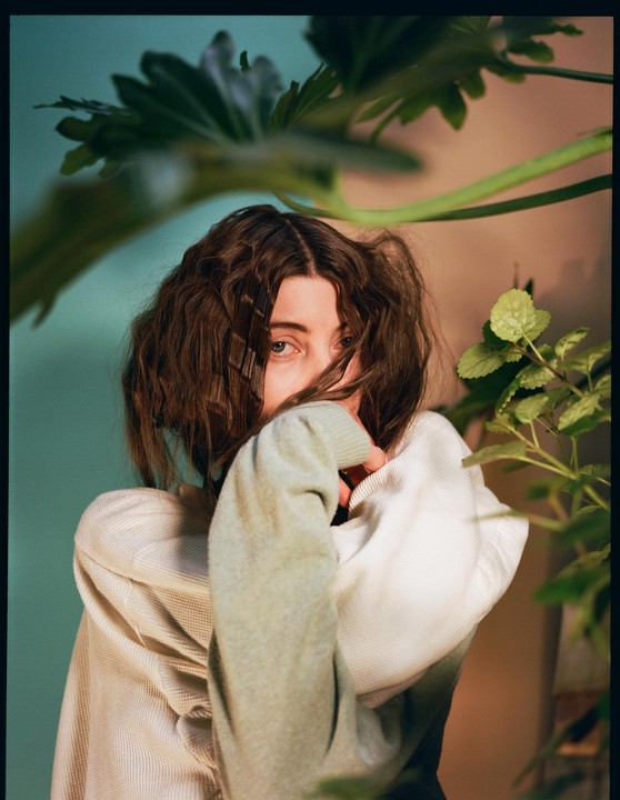 A woman with curly hair looks directly at the viewer, partially obscured by her arm and green foliage.