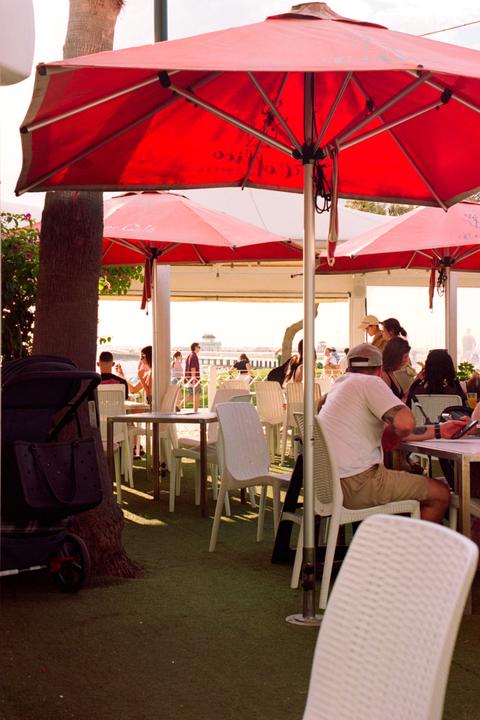 A cafe patio with red umbrellas, white chairs, and people sitting at tables.