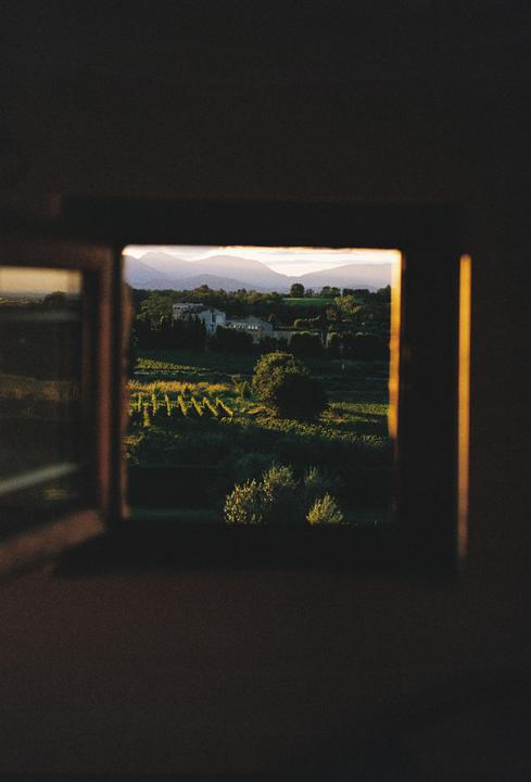 A view of a vineyard, buildings, and mountains is seen through a window frame at sunrise.