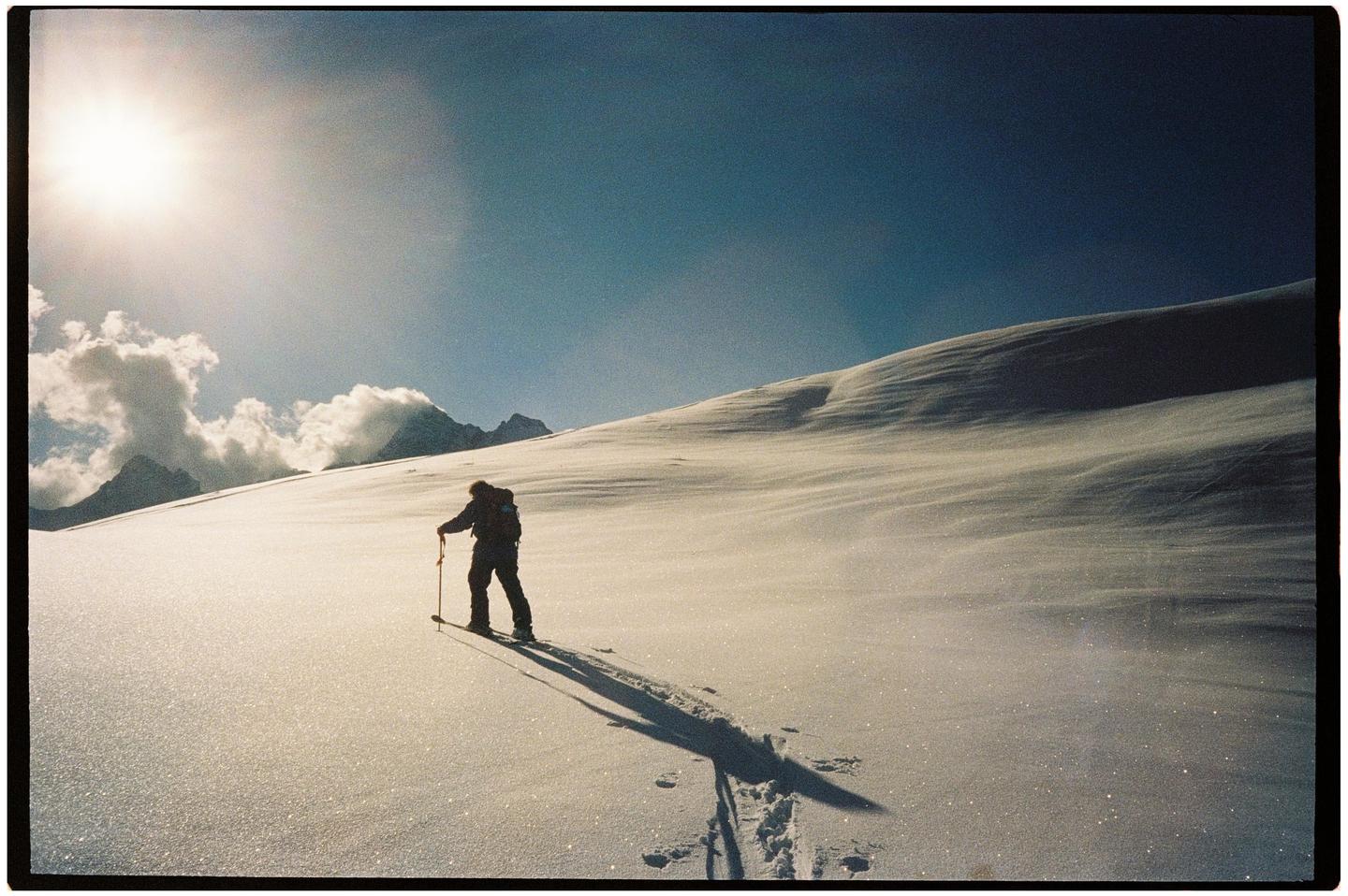 A person on skis with poles is silhouetted against a bright sky and snow-covered mountain.