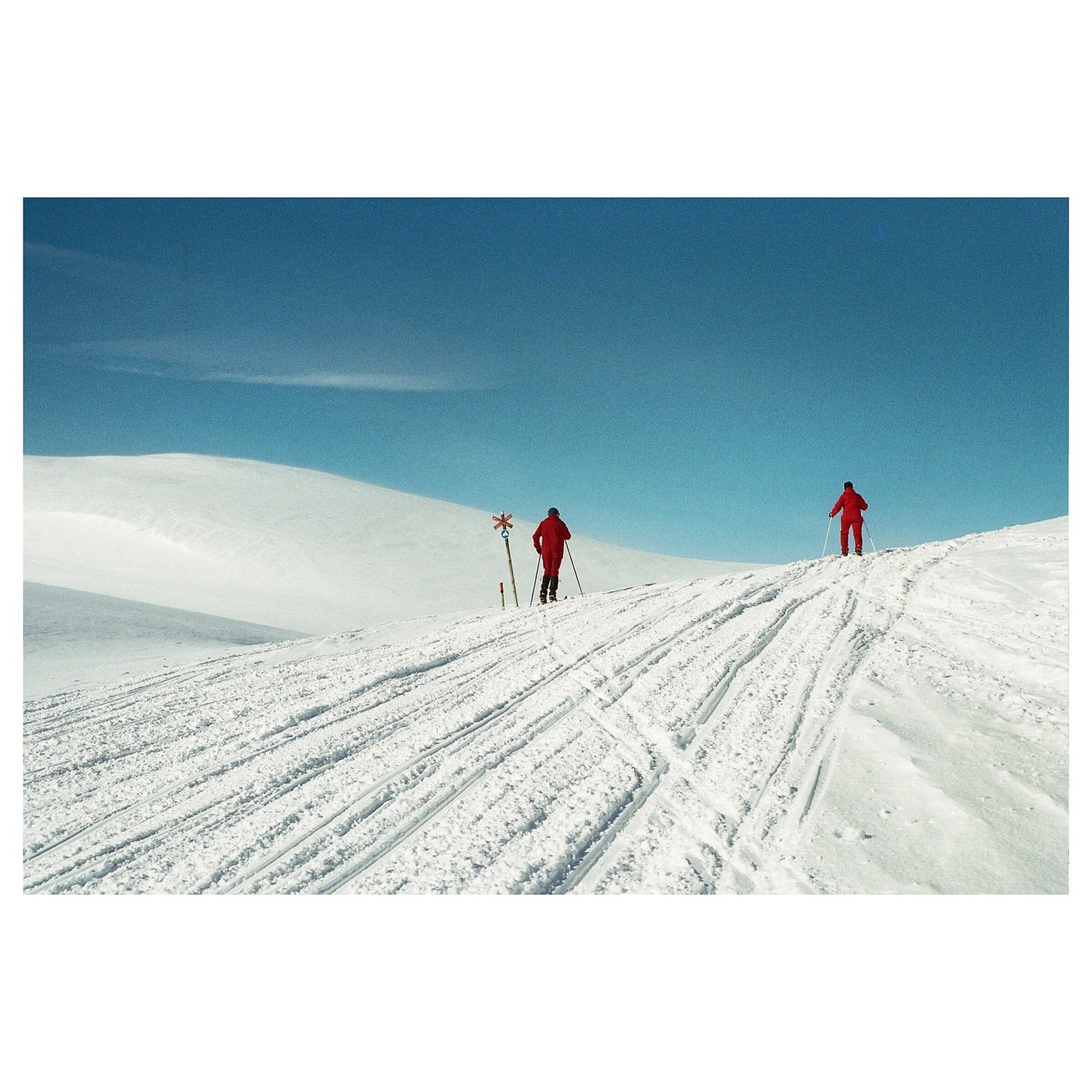 Two people in red ski suits are cross-country skiing up a snowy hill under a clear blue sky.