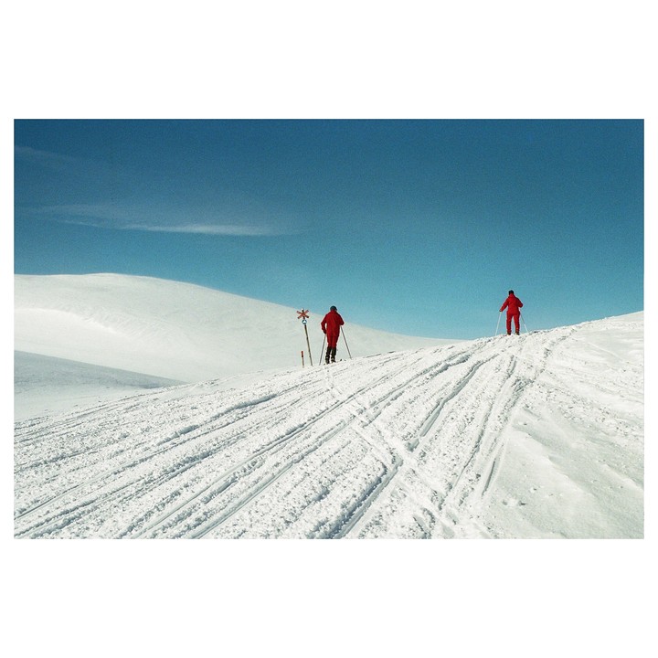 Two people in red ski suits are cross-country skiing up a snowy hill under a clear blue sky.