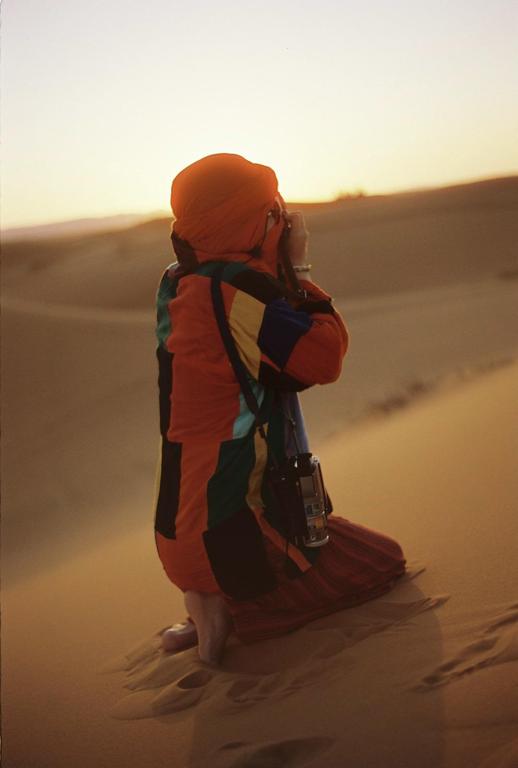 A person in colorful clothing and a head covering kneels in a desert, holding a camera to their eye at sunset.