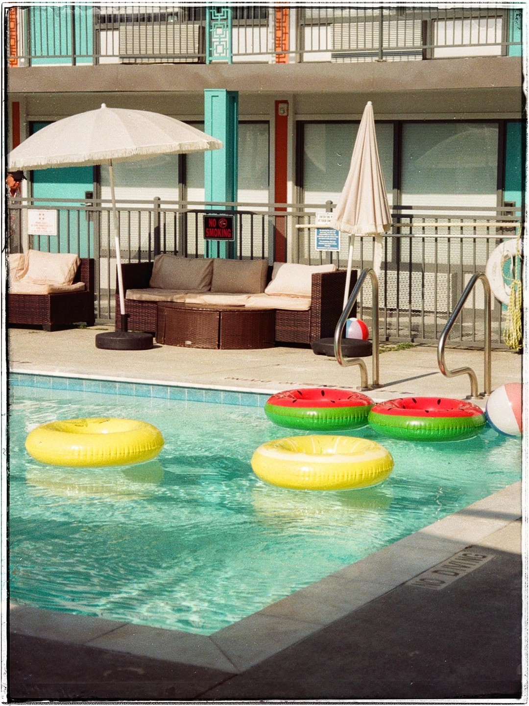 Several inflatable rings float in a swimming pool next to a patio with outdoor furniture and a building.