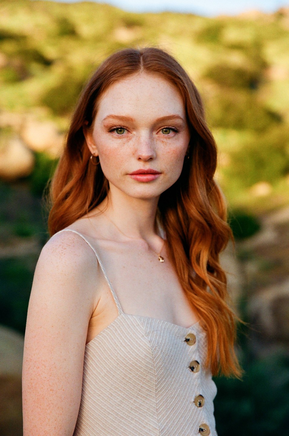 A woman with long red hair and freckles looks directly at the viewer in a portrait taken outdoors.